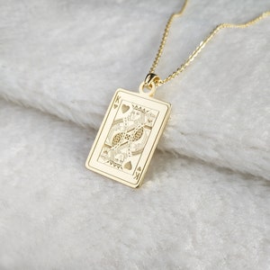 May include: A gold pendant necklace. The pendant is a rectangular shape, depicting the King of Hearts playing card. The card design includes the suit symbols and the letter "K". The pendant hangs from a fine gold chain. The necklace is a stylish accessory.