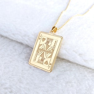 May include: A gold pendant necklace with a playing card design. The pendant is rectangular and depicts the Jack of Hearts card. The necklace has a fine chain and a clasp. The pendant is made of a shiny, reflective material, and is suspended from a delicate chain.