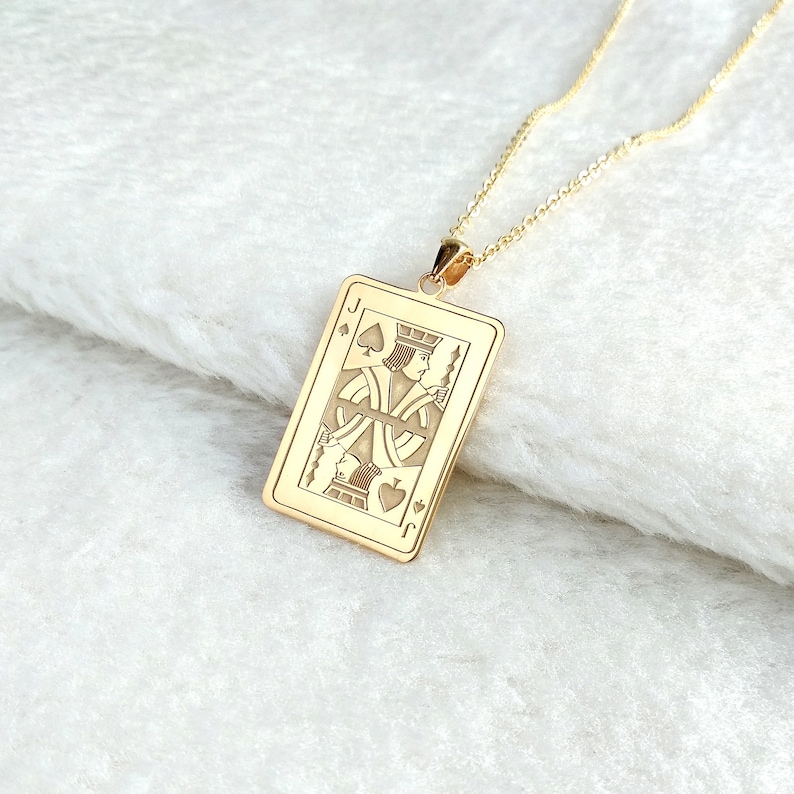 May include: Gold-coloured pendant necklace featuring a rectangular pendant with a detailed engraving of the Jack of Spades playing card. The pendant hangs from a delicate gold chain, set against a white, textured surface.