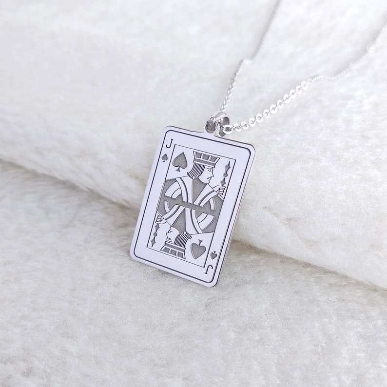 May include: A silver pendant necklace with a detailed engraving of the Jack of Spades playing card. The rectangular pendant with rounded corners hangs from a delicate chain. The card design includes the spade symbol and the letter "J".