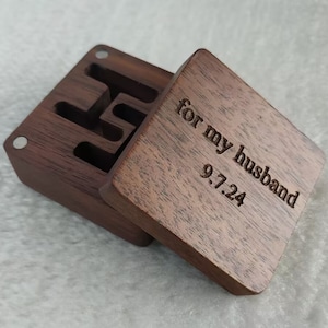 May include: A dark brown wooden box with a hinged lid. The lid is open and the inside of the box is visible. The lid is engraved with the text "for my husband 9.7.24".