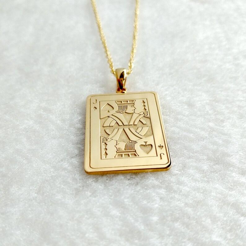 May include: Gold pendant necklace. The rectangular pendant features an engraved Jack of Spades playing card design. The pendant is suspended from a fine gold chain. The card design includes the letter "J" and the spade symbol.