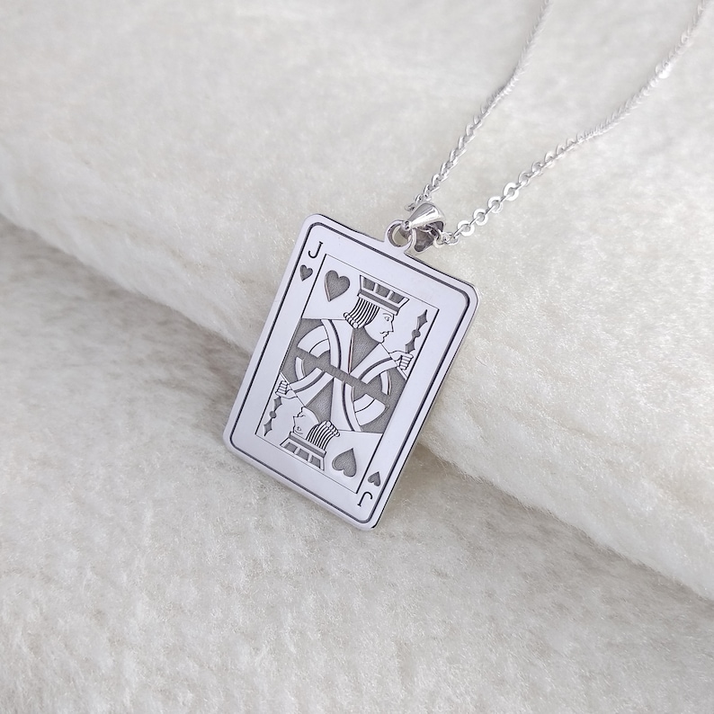 May include: Silver pendant necklace. The pendant is rectangular and features the Jack of Hearts playing card design. The card's details are engraved, including suit symbols and the letter "J". The necklace chain is also silver, creating a cohesive look.