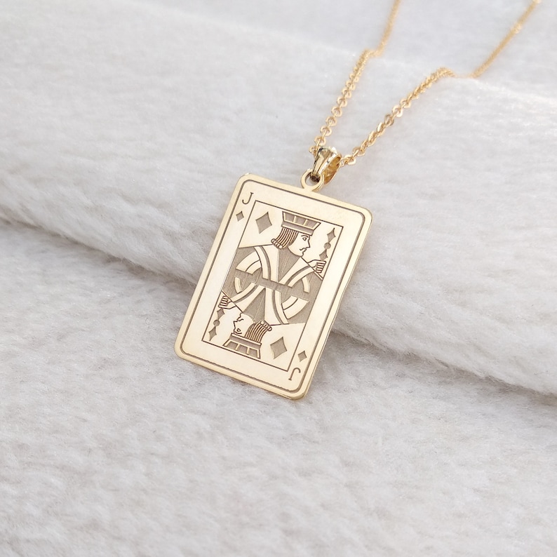May include: A gold-coloured pendant necklace. The pendant is a rectangular shape, resembling a playing card, and features the Jack of Diamonds. The card design is intricately engraved. The necklace chain is also gold-coloured.