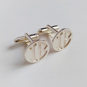 Puede incluir: Un par de gemelos de plata con un dise&ntilde;o de monograma. Los gemelos son de forma ovalada y presentan las letras "IB" en una fuente en negrita sin serif.