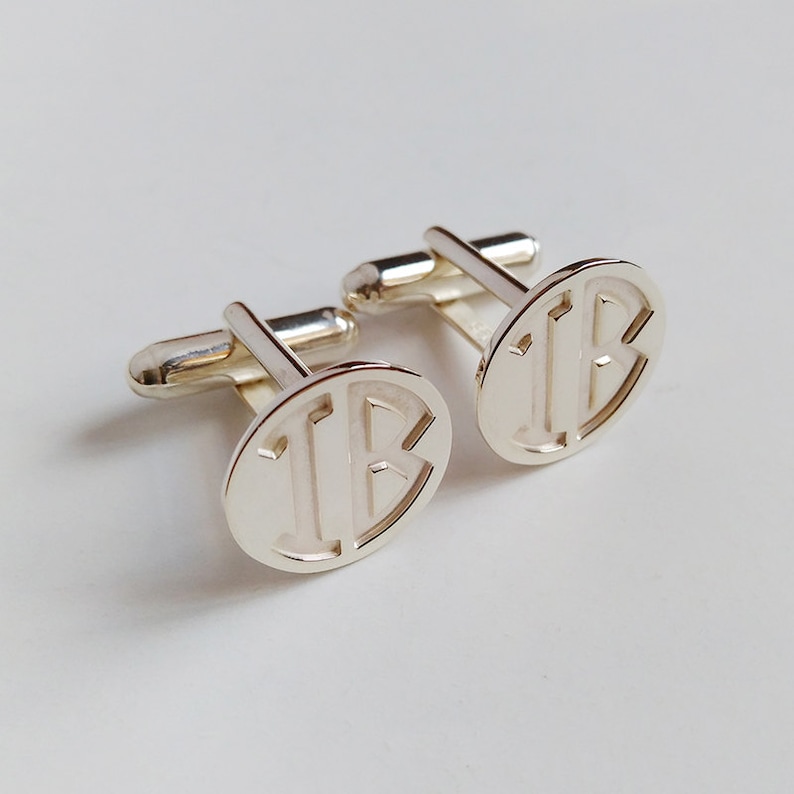 Puede incluir: Un par de gemelos de plata con un dise&ntilde;o de monograma. Los gemelos son de forma ovalada y presentan las letras "IB" en una fuente en negrita sin serif.