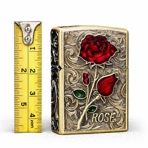 Op de afbeelding: Een messingkleurige aansteker met een rood rozenontwerp. De aansteker heeft een sierlijk, gegraveerd patroon en het woord "ROSE" staat op de voorkant. De aansteker is ongeveer 5 cm hoog.