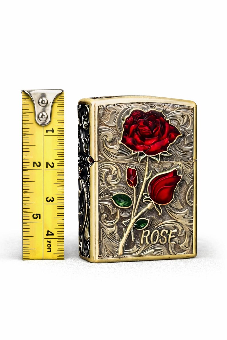 Op de afbeelding: Een messingkleurige aansteker met een rood rozenontwerp. De aansteker heeft een sierlijk, gegraveerd patroon en het woord "ROSE" staat op de voorkant. De aansteker is ongeveer 5 cm hoog.