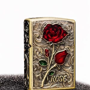 Op de afbeelding: Een vintage aansteker met een gedetailleerd bloemmotief. De aansteker heeft een rode roos met groene bladeren en het woord "ROSE" op de voorkant. Het metaal heeft een goud- en zilverafwerking.