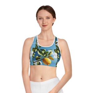 May include: A light blue sports bra featuring a vibrant lemon tree design. The bra showcases a sunny sky background with lemons, leaves, and branches. The bra has a white trim and a racerback style.