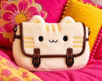 May include: A cream-colored plush cat-shaped crossbody bag with soft faux fur texture. The bag features embroidered yellow stripes, black circular eyes, a small black mouth, and two small ears. It has brown faux leather trim and two buckles.