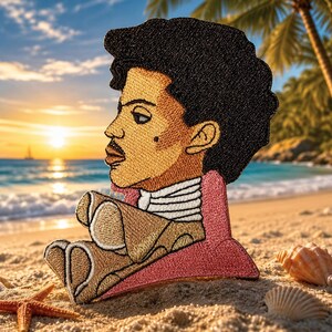 May include: Embroidered patch featuring a profile portrait of a person with dark curly hair, a mustache, and a beauty mark. The patch is in shades of tan, black, and pink, with a white collar. The patch is sitting on a sandy beach with a sunset in the background.