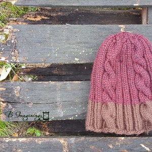 May include: A knitted hat with a cable knit pattern in burgundy and brown. The hat is lying on a wooden surface.