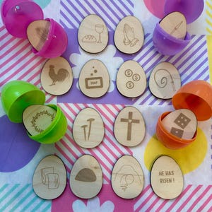 Puede incluir: Adornos de huevo de Pascua de madera con símbolos religiosos grabados y texto, incluyendo una cruz, clavos y la frase "HE HAS RISEN!". Algunos huevos están dentro de huevos de Pascua de plástico de colores. El fondo es una tela estampada colorida.