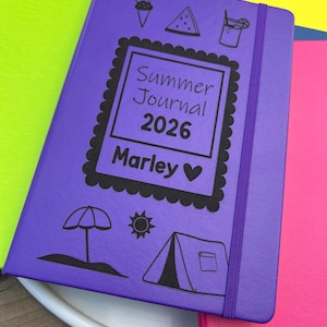 以下が含まれることがあります： 「Summer Journal 2026 Marley」の文字と夏のイラストが描かれた紫色のジャーナル。ジャーナルにはお揃いのゴムバンド留めがあり、他のカラフルなノートに囲まれています。