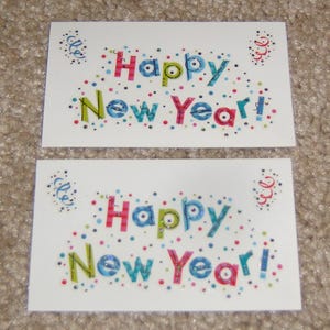 May include: Two white greeting cards with the text "Happy New Year!" in colorful, glittery letters. The cards are decorated with confetti and swirls. The background is a neutral, textured surface.