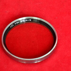 May include: Close-up view of a silver-rimmed filter for cameras. The filter is circular and features the text 'B + W 52E KR3 12X' printed on its rim. The filter's glass is clear and sits within the metallic frame. The filter is displayed against a red fabric background.