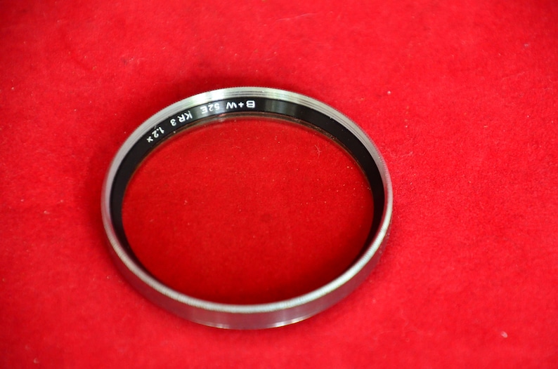 May include: Close-up view of a silver-rimmed filter for cameras. The filter is circular and features the text 'B + W 52E KR3 12X' printed on its rim. The filter's glass is clear and sits within the metallic frame. The filter is displayed against a red fabric background.