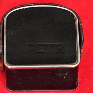 May include: Close-up view of a vintage Petri camera case. The case is black leather with a silver metal frame and the word 'PETRI' embossed on the front.  The case is rectangular with a slightly curved top. The leather shows some signs of wear.  It's a compact, classic camera accessory, ideal for collectors or photography enthusiasts.