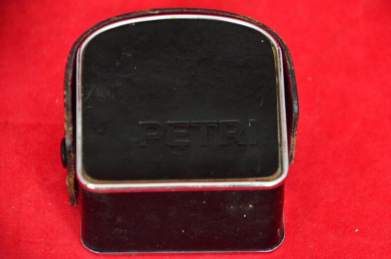 May include: Close-up view of a vintage Petri camera case. The case is black leather with a silver metal frame and the word 'PETRI' embossed on the front.  The case is rectangular with a slightly curved top. The leather shows some signs of wear.  It's a compact, classic camera accessory, ideal for collectors or photography enthusiasts.