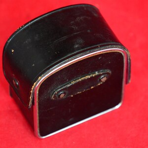 May include: Vintage black leather camera case with silver metal trim. The case has a rectangular shape with a slightly curved top.  The leather shows some signs of wear and discoloration.  A small leather strap with metal rivets is visible on the back. The case is ideal for storing and protecting a vintage camera or other small device.