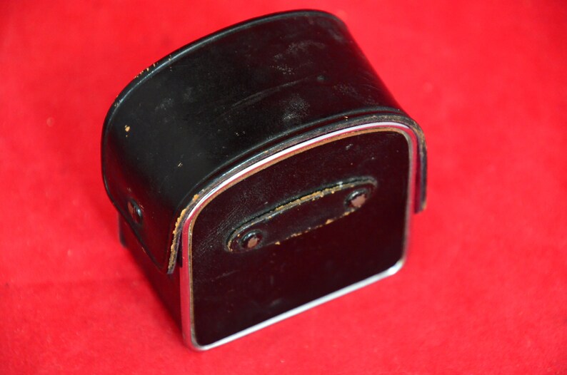 May include: Vintage black leather camera case with silver metal trim. The case has a rectangular shape with a slightly curved top.  The leather shows some signs of wear and discoloration.  A small leather strap with metal rivets is visible on the back. The case is ideal for storing and protecting a vintage camera or other small device.