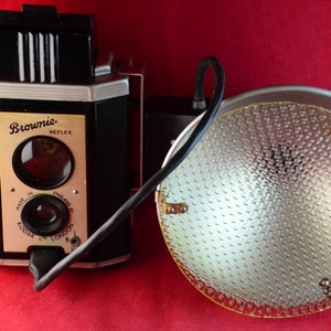 May include: A black and silver Brownie Reflex camera with a flash attachment. The camera has a viewfinder and a lens. The flash attachment is connected to the camera by a black cord.