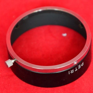 May include: Close-up view of a black lens hood with a silver metallic rim. The lens hood has a small, silver adjustment screw on the side and the word 'PETRI' is visible on its surface. The lens hood is circular and appears to be made of metal and plastic.  It's shown against a solid red background.