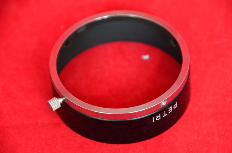 May include: Close-up view of a black lens hood with a silver metallic rim. The lens hood has a small, silver adjustment screw on the side and the word 'PETRI' is visible on its surface. The lens hood is circular and appears to be made of metal and plastic.  It's shown against a solid red background.