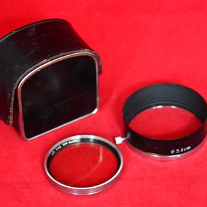May include: Close-up view of camera accessories on a red surface.  A black leather camera case with a metallic trim is shown.  Also included are a lens hood with Ø5.4cm marking and a filter, both with metallic rims. The accessories appear vintage or classic in style.