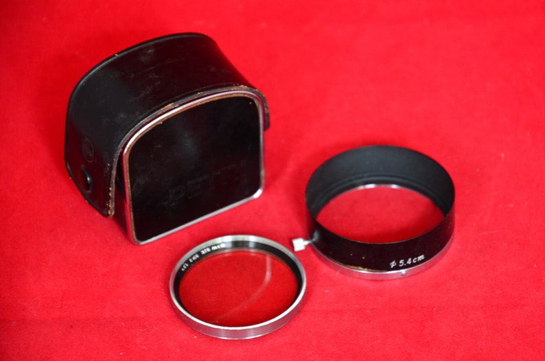 May include: Close-up view of camera accessories on a red surface.  A black leather camera case with a metallic trim is shown.  Also included are a lens hood with Ø5.4cm marking and a filter, both with metallic rims. The accessories appear vintage or classic in style.