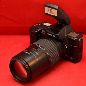 May include: A black 35mm film camera with a built-in flash and a black Canon lens attached. The camera has a silver label that reads "Chinon" and the lens has a silver label that reads "Canon FD 100-300mm 1:5.6".