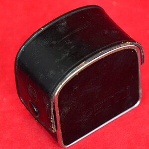 May include: Close-up view of a vintage black leather camera case with a silver metallic trim. The case shows signs of wear and is slightly scuffed.  The word 'Pentax' is subtly embossed on the front. The case is rectangular with a curved top.  The overall condition suggests age and use, characteristic of vintage camera accessories.