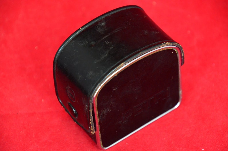 May include: Close-up view of a vintage black leather camera case with a silver metallic trim. The case shows signs of wear and is slightly scuffed.  The word 'Pentax' is subtly embossed on the front. The case is rectangular with a curved top.  The overall condition suggests age and use, characteristic of vintage camera accessories.