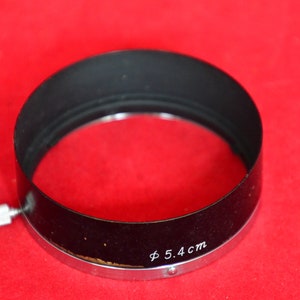 May include: Close-up view of a black lens hood with a silver metal rim. The lens hood is circular with a slight curve and features a small, silver screw-on attachment.  The text 'Φ 5.4 cm' is inscribed on the hood. The lens hood is displayed against a solid red background.