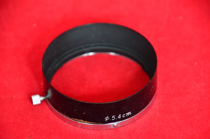 May include: Close-up view of a black lens hood with a silver metal rim. The lens hood is circular with a slight curve and features a small, silver screw-on attachment.  The text 'Φ 5.4 cm' is inscribed on the hood. The lens hood is displayed against a solid red background.