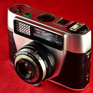 Peut inclure: Un appareil photo argentique 35 mm argenté et noir avec un revêtement en simili cuir noir. L'appareil photo a un objectif avec une bague noire et argentée et un dessus argenté avec un flash intégré. L'appareil photo porte l'inscription "Revue 300 CS" sur le dessus.