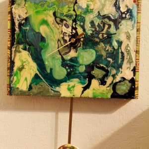 May include: A green, blue, and white abstract clock with a gold pendulum. The clock face is made of a repurposed cigar box.