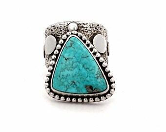 May include: A ring featuring a triangular turquoise stone with dark veining, set in a silver-toned metal band. The band has a textured, pitted surface and is accented with smooth, circular silver-toned discs on the sides and a beaded border.