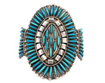 May include: A silver cuff bracelet with an oval turquoise stone design. The bracelet features numerous small turquoise stones set in a silver frame. The design is symmetrical and detailed, showcasing a Native American jewelry style.