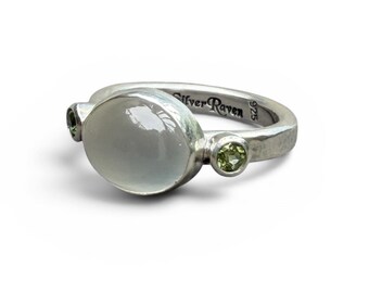 May include: A sterling silver ring featuring a large, smooth, oval-shaped white moonstone cabochon flanked by two small, round, faceted green peridot gemstones. The inner band is engraved with the text Silver Raven and the hallmark 925.
