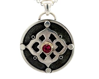 May include: Silver pendant necklace featuring a circular pendant with a textured black background and a detailed silver overlay. The overlay has an intricate design with a central red gemstone. The necklace chain is also silver.
