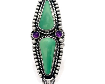 May include: A silver ring with two green turquoise stones and two purple amethyst stones. The ring has a detailed design with a rope-like pattern.