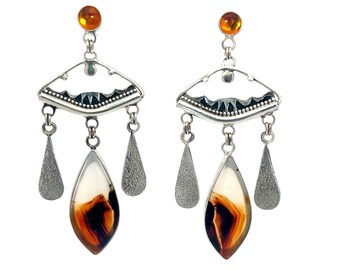May include: A pair of silver dangle earrings with a teardrop-shaped agate stone in the center of each earring. The agate stones are brown and white with a marbled pattern. The earrings have a silver arch with a decorative design above the stones and three silver teardrop-shaped charms hanging from the arch.