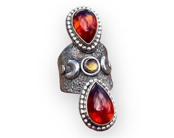 May include: A silver ring with three red and orange teardrop-shaped stones. The ring has a textured band with a crescent moon design.