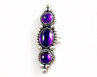 May include: A silver ring with three purple amethyst stones set in a bezel setting. The stones are surrounded by a decorative silver border.