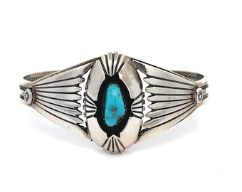 May include: A silver cuff bracelet with a central turquoise stone. The bracelet features a detailed design with radiating lines and decorative elements. The turquoise stone is oval-shaped and set in a black bezel. The bracelet is displayed on a white background.