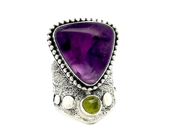 May include: A silver ring with a triangular amethyst gemstone and two smaller green gemstones. The ring has a textured band.