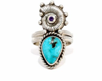 May include: A silver ring featuring a teardrop-shaped turquoise stone with black matrix, bordered by a twisted silver wire. Above the stone sits a silver ammonite fossil motif accented with a small, round, purple amethyst gemstone at its center.