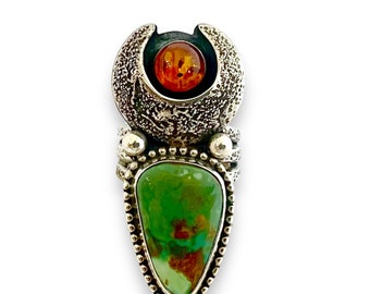 May include: A handcrafted pendant featuring a vibrant green turquoise stone and a round amber cabochon. The pendant has a textured silver setting with a unique design. The turquoise stone is teardrop-shaped, and the amber is set in a textured, circular frame.